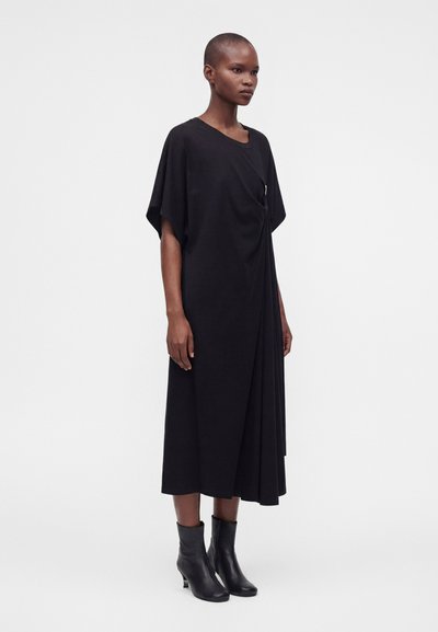 Femme debout portant une robe midi noire ample à manches courtes et des bottines noires sur un fond clair uni.