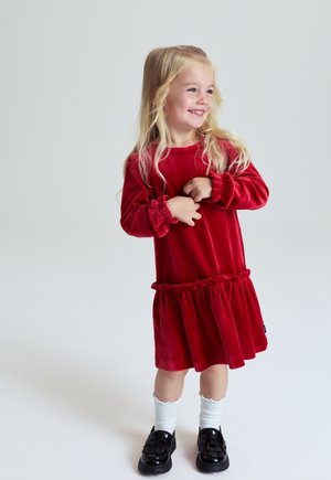 Junges Mädchen mit langen blonden Haaren, das lächelt. Sie trägt ein rotes Samtkleid, weiße Socken und schwarze Schuhe und steht vor einem schlichten Hintergrund.