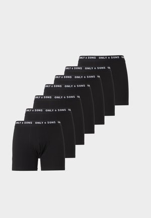 Homme athlétique debout pieds nus, portant un boxer noir avec une ceinture de marque, regardant vers sa gauche sur un fond clair uni.