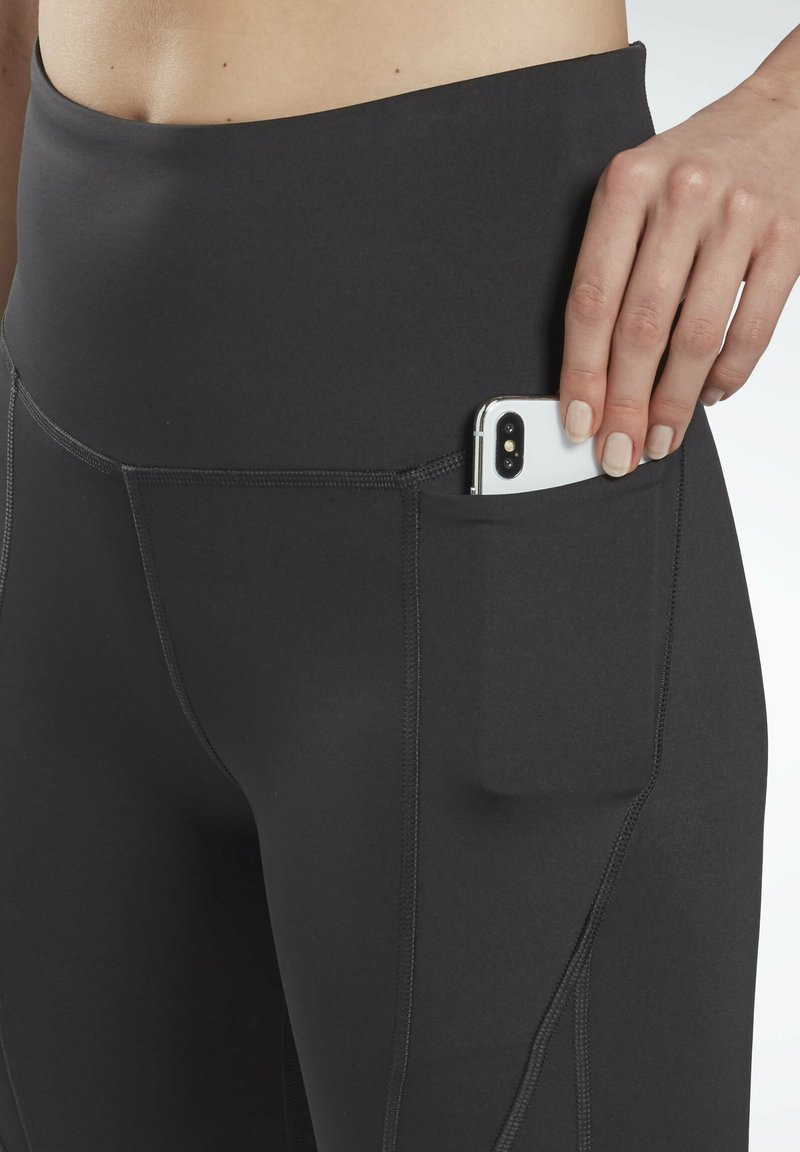 Leggings de sport noirs avec une taille haute, dotés d'une poche latérale contenant un smartphone. Texture lisse avec des coutures contrastées.