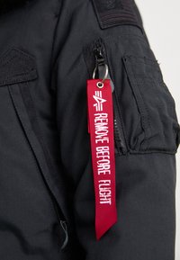Čierna izolačná bunda s vreckom na zips, nášivkou americkej vlajky a červeným štítkom "REMOVE BEFORE FLIGHT" pripevneným na kľúčenke.