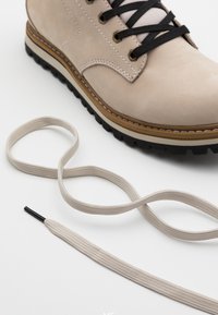 Botte en daim beige avec des lacets noirs, un bout arrondi, et une semelle en caoutchouc marron. Comprend un lacet plat beige clair à proximité.