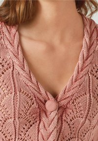 Primo piano di una donna che indossa un cardigan lavorato a maglia rosa con un motivo a foglia testurizzato e un bottone rivestito di tessuto sul colletto.