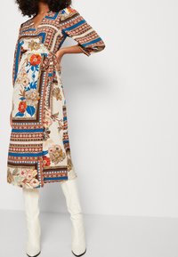 Romantische wikkeljurk met bloemenpatroon in crème, met blauwe en rode geometrische accenten, driekwart mouwen en een vastgebonden taille. Gecombineerd met witte kniehoge laarzen.