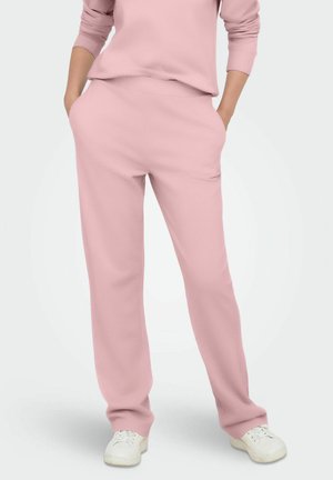 Vrouw in lichtroze casual broek en bijpassend topje met handen in de zakken en witte sneakers, staande op een lichte achtergrond.