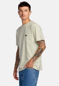 T-shirt verde chiaro a maniche corte con tasca e un logo nero. Chi lo indossa ha tatuaggi visibili e lo abbina a jeans blu.