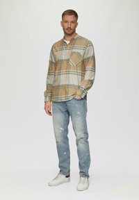 Knoop-up geruite shirt in beige, wit en blauw; enkel borstzakje. Lichtblauwe versleten jeans met verfspetters. Witte sneakers.