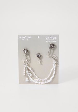 Charm Jibbitz Crocs con hardware argentato, perle bianche e spilli di sicurezza su uno sfondo grigio con testo del marchio e la nota "Unicamente Te."