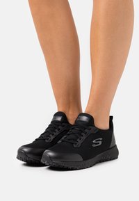 Schwarze Sportschuhe mit einem Obermaterial aus Mesh, einer strukturierten Gummisohle und tonal abgestimmtem Branding an der Seite und der Zunge. Sie verfügen über Schnürsenkel und ein rutschfestes Design.