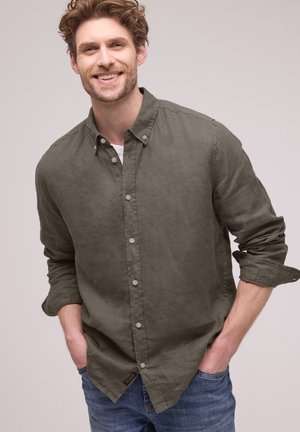 Smilende mand med krøllet hår iført olivengrøn knap-op-skjorte med opkørte ærmer og blå jeans, stående med hænderne i lommerne.
