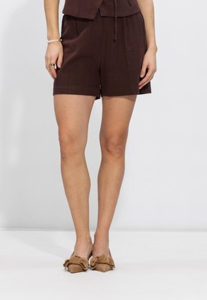 Vrouw draagt donkerbruine korte broek en beige puntige platte schoenen met grote strikken, staand tegen een effen lichte achtergrond.