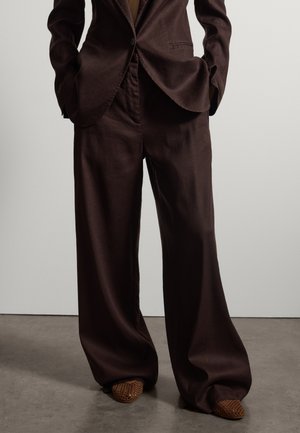 Personne portant un pantalon ample marron foncé à jambes larges et un blazer assorti, debout les mains dans les poches, portant des chaussures tissées marron.