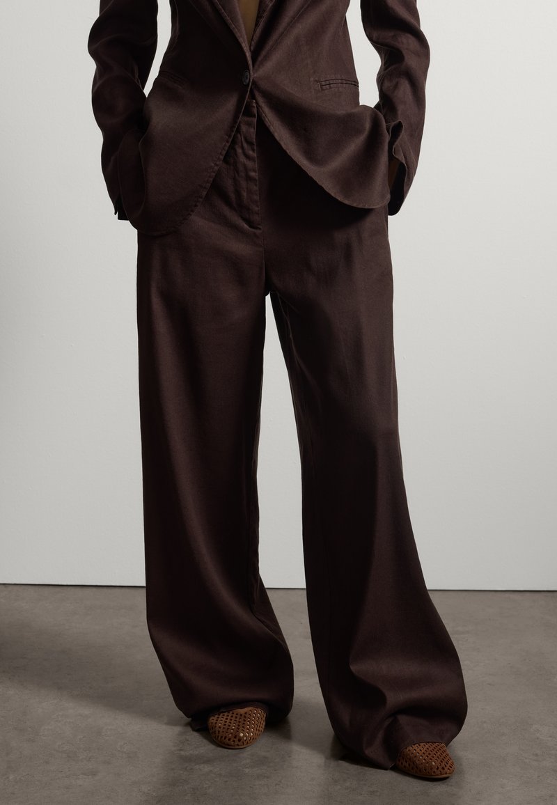 Person trägt locker sitzende dunkelbraune Hosen mit weitem Bein und passendem Blazer, steht mit den Händen in den Taschen und trägt gewebte braune Schuhe.