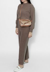 Tuta marrone a maglia con maniche lunghe, abbinato a una borsa a spalla metallica color rosa dorato. Le scarpe bianche slip-on completano l'outfit.