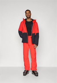 Ulkoilulaskettelutakki punaisena ja tummansinisenä, jossa on huppu, vetoketjullinen suljetus ja joustavat hihansuut, yhdistettynä sopiviin punaisiin housuihin ja laskettelukenkiin.