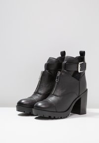 Czarne skórzane botki za kostkę na wysokim słupku, zapinane na zamek błyskawiczny, z akcentem w postaci klamry. Teksturowane wykończenie z rowkowaną gumową podeszwą dla lepszej przyczepności.