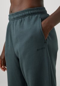 Sötétzöld melegítőnadrág, elasztikus derékkal és oldalsó zsebekkel, puha anyagból, oldalt diszkrét logóval díszítve.