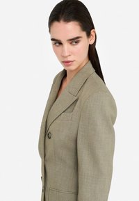 Blazer su misura verde oliva con cuciture sulle spalle strutturate, chiusura con un bottone e due tasche frontali. Tessuto testurizzato con un motivo sottile.