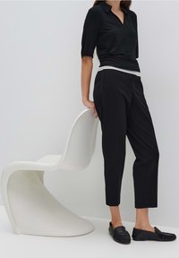 Femme portant un pantalon noir court, un haut noir, des mocassins noirs, debout à côté d'une chaise blanche moderne et incurvée sur un fond uni.
