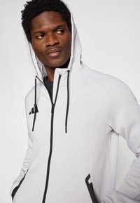 Φωτεινή γκρι ζακέτα με φερμουάρ, που διαθέτει μαύρο λογότυπο, ρυθμιζόμενα κορδόνια και υφασμάτινες λεπτομέρειες στους ώμους. Μαλακό ύφασμα, αθλητική εφαρμογή.