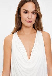 Bílý top bez rukávů s hlubokým véčkovým výstřihem a drapovaným designem látky. Měkká textura a volný střih zdůrazňují plynulou siluetu.