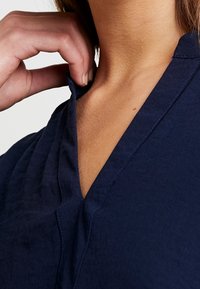 Haut bleu marine avec un col en V, fabriqué en tissu texturé. Une main saisit le col, mettant en valeur son détail décontracté.