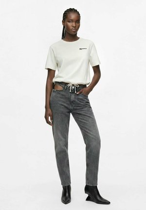 Donna nera con camicia bianca a maniche corte, jeans grigi a vita alta con ritagli e stivaletti neri, in piedi con una mano in tasca.