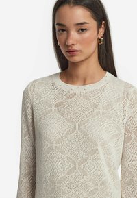 Stickad krämfärgad tröja med diamantsymptom, rund halsringning och långa ärmar. Det lätta materialet har en öppen vävstruktur.