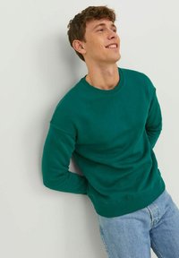 Felpa girocollo verde in una morbida miscela di cotone, con polsini e orlo a coste, indossata con jeans in denim azzurro chiaro. Design semplice e minimal.