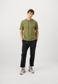 Olivgrünes T-Shirt mit kurzen Ärmeln, kombiniert mit schwarzen Hosen und beigen Sneakers. Minimalistisches Design, normale Passform, Baumwollmischgewebe.