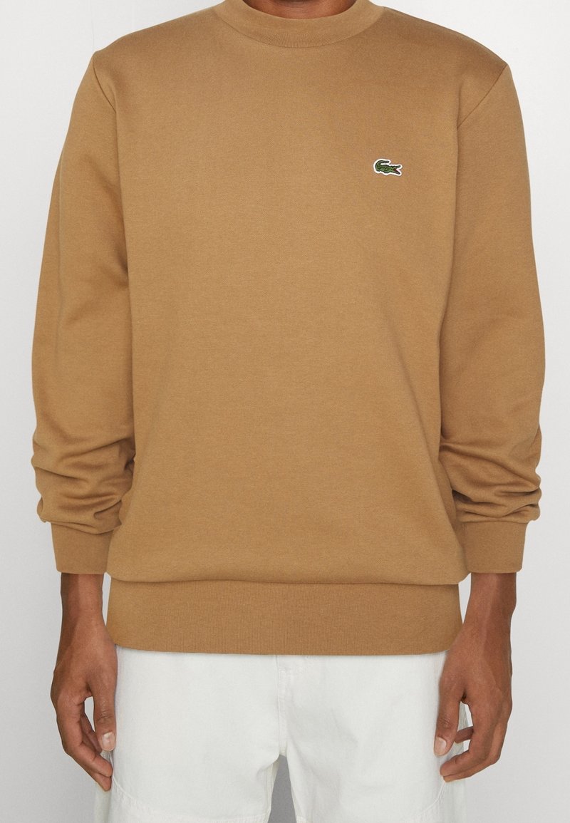 Sudadera marrón de algodón con mangas largas, cuello redondo acanalado y un pequeño logo verde de Lacoste en el pecho. Textura suave, corte relajado.