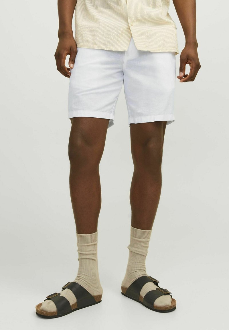 Pantalones cortos de lino blancos con puños enrollados, combinados con sandalias negras y beige y calcetines beige hasta la rodilla. Enfocado en un estilo casual de verano.