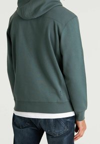 Felpa con cappuccio di un verde tenue, realizzata in tessuto morbido. Presenta maniche inserite e dettagli testurizzati lungo il retro. Abbinata a jeans scuri.