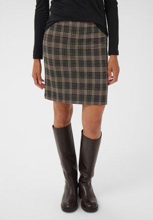 Femme portant une jupe longue marron à carreaux jusqu’aux genoux, un haut noir à manches longues et des bottes en cuir marron foncé montant jusqu’aux genoux, debout sur fond blanc.