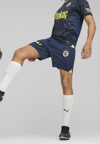 Sininen jalkapallovarustus, jossa on kamouflagekuvio, keltaiset yksityiskohdat ja logo. Valkoiset sukat ja mustat nappulakengät täydentävät asun.