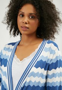 Cardigan blu con motivo a zigzag e accenti bianchi, caratterizzato da scollo a V e bordi a coste. Indossato sopra un top in pizzo bianco.