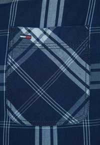 Látka námořnické kostkované košile s náprsní kapsou, která má kontrastní prošívání a červenobílo-modrý logo detail na švu kapsy.