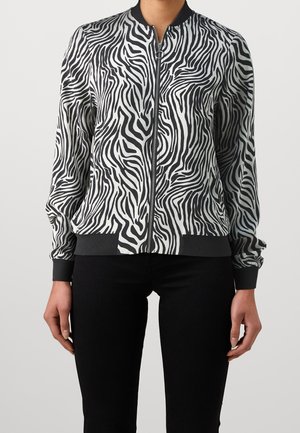 Kvinde iført en sort-hvid zebra-stribet lynlåstrøje med ribkanter ved ærmer og kant, kombineret med sorte bukser, mod en ensfarvet baggrund.