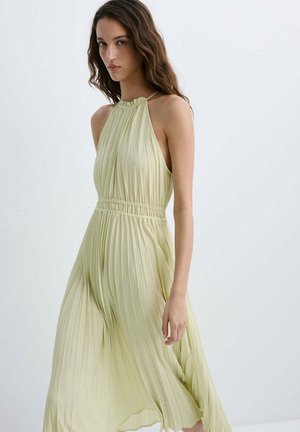 Femme portant une robe à dos nu plissée vert clair, debout devant un fond blanc uni.