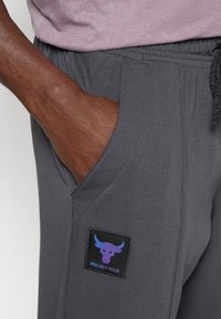 Graue Sporthosen mit einer Seitentasche, die ein schwarzes Patch mit einem lila Stier-Logo und dem Text "PROJECT ROCK" zeigen. Weicher, flexibler Stoff.