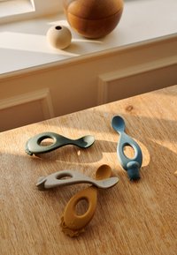 Four assorted silicone spoons in green, blue, beige, and yellow, featuring playful designs with oval cutouts and textured ends, on a wooden surface.