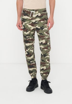 Mann trägt grüne, braune und beige Camouflage-Cargohose mit elastischen Bündchen und schwarze Sneaker, steht vor weißem Hintergrund.