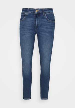 Jean bleu ajusté en denim avec une taille mi-haute, présentant cinq poches et des détails de coutures subtiles, porté avec des baskets noires.