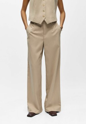 Femme portant un pantalon large beige à rayures fines avec un gilet boutonné assorti, mains dans les poches, et chaussures foncées à bout carré.