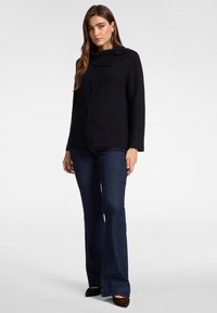 Vrouw die een zwarte geribbelde trui met een opgevouwen kraag, donkerblauwe uitlopende jeans en bruine puntige schoenen draagt, staand tegen een effen achtergrond.