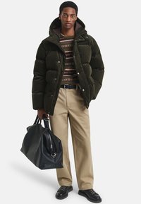 Giacca a vento verde con cappuccio, maglione a fantasia sotto, pantaloni beige chiaro e una borsa a tracollo nera in pelle. Scarpa nera completa il look.