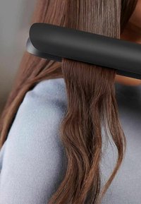 Un fer à lisser au design noir élégant, lissant des cheveux bruns ondulés. Les cheveux brillent avec une texture variée, sur fond de pull bleu clair.
