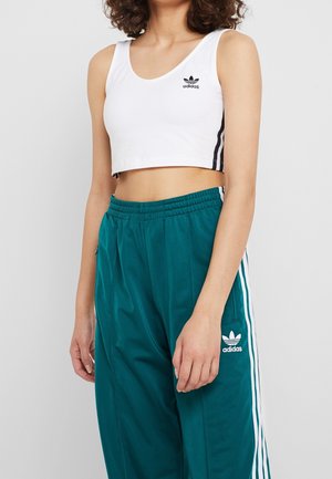 Vrouw draagt een wit Adidas cropped tanktop en teal Adidas trainingsbroek met witte strepen, staand tegen een lichte achtergrond.