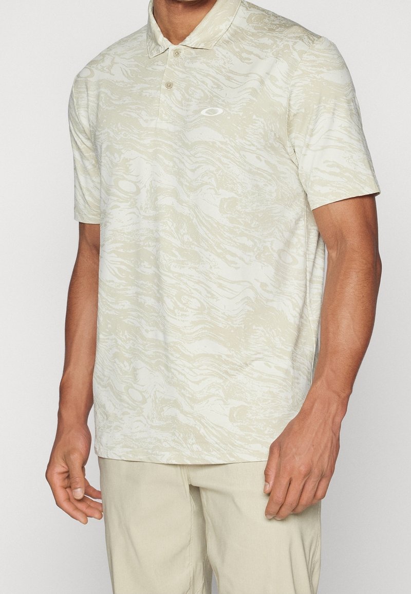 Homme portant un polo beige à manches courtes avec un motif ondulé subtil et un petit logo sur la poitrine, associé à un pantalon beige clair.