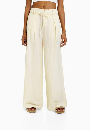 Krämfärgade vida byxor med dragsko i midjan, plisserad design och mjuk textur, tillsammans med bruna plattformssandaler.